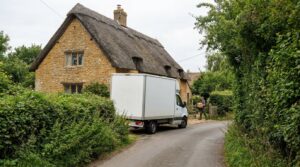 A plain removal van on a narrow lane beside a thatched stone cottage, with a mover carrying a box through the gate